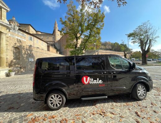 Noleggio Vitale NCC Basilicata: un elegante van nero con il logo Vitale NCC parcheggiato su una strada di ciottoli, con un edificio storico sullo sfondo.