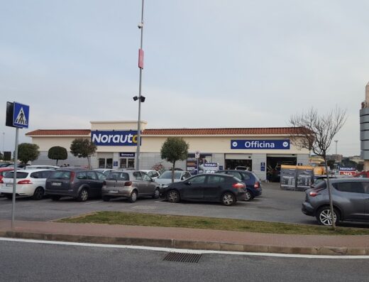 Norauto Fiumicino: vista esterna del negozio con parcheggio auto, insegne "Norauto" e "Officina", e torre cilindrica sullo sfondo. Centro specializzato in accessori e servizi per auto e moto.