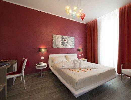 Accogliente camera dell'Oasi Village Hotel con letto matrimoniale decorato con cigni di asciugamano e petali, pareti bordeaux, scrivania e poltrona bianca.