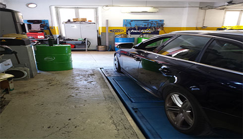 Interno dell'officina MT Autoservice a Genova con auto nera in riparazione su piattaforma elevatrice, barili di olio motore Ford e attrezzature per la manutenzione auto.