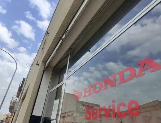 Officina Autorizzata Honda a Trapani, Italia. Vetro con insegna rossa Honda Service, riflessi del cielo e degli edifici circostanti.