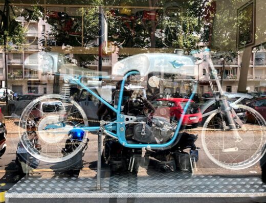 Esposizione di una motocicletta d'epoca blu presso Officina Colognese, con riflessi urbani sulla vetrina. Ricambi e accessori moto visibili.