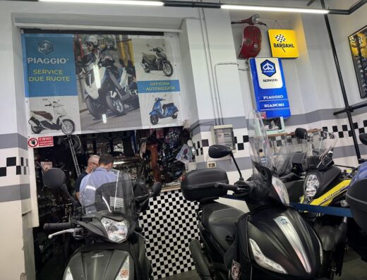 Interno di Officina Due Ruote con scooter Piaggio in primo piano, insegne Bardahl e cartello Piaggio Service. Riparazione e vendita accessori moto.