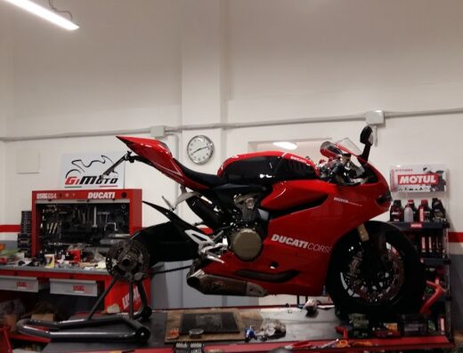 Officina GiMoto: Ducati Corse rossa sollevata per manutenzione, senza ruota anteriore. Attrezzi e logo GiMoto visibili. Servizio di riparazione moto.