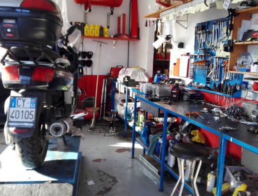 Officina MeccanoMoto: scooter con bauletto in riparazione, meccanico al lavoro, attrezzi e banco da lavoro visibili. Servizi di riparazione moto.