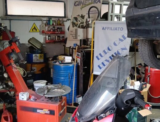 Interno dell'officina moto Ginestra, con una moto rossa in primo piano, attrezzature per la riparazione e una vettura sollevata. Servizi di revisione e riparazione moto.