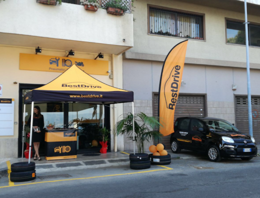 Officina Musumeci Service: esterni dell'officina con tendone e bandiera BestDrive, auto nera parcheggiata, pneumatici e hostess per la revisione auto.