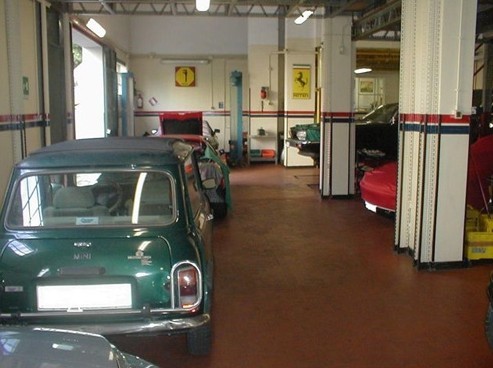 Interno dell'Officina New Oram con Mini Cooper verde in primo piano. Si vedono altre auto, Ferrari e attrezzature da officina.
