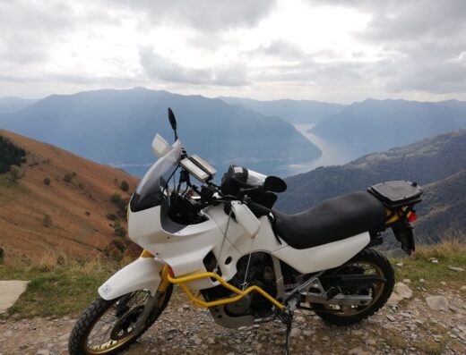 Moto da turismo adventure bianca parcheggiata con vista panoramica sulle montagne e sul lago. Accessori da viaggio e protezioni gialle.