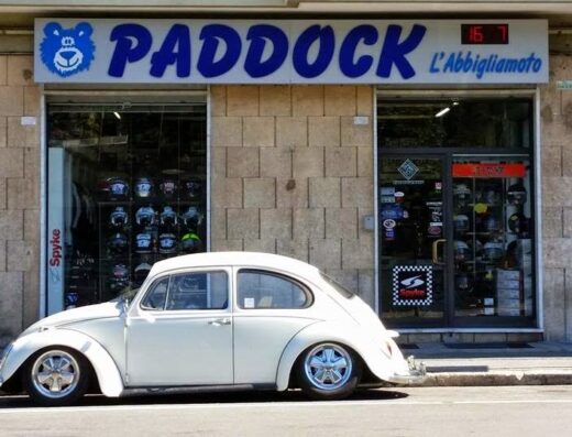 Auto d'epoca bianca parcheggiata di fronte al negozio Paddock L'Abbigliamoto, specializzato in accessori e abbigliamento per motociclisti. In vetrina caschi moto e altri prodotti.