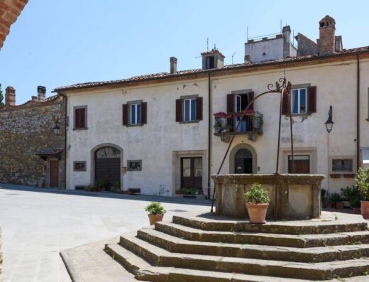 Palazzo Ninci: View of a weathered stone well in a piazza with cream-colored buildings, brown shutters, and potted plants under a bright sky in Tuscany, Italy.