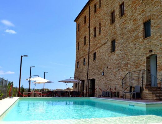 Palazzo Riccucci Resort Montappone swimming pool, featuring a clear blue pool, umbrellas, tables and chairs, and a brick building.
