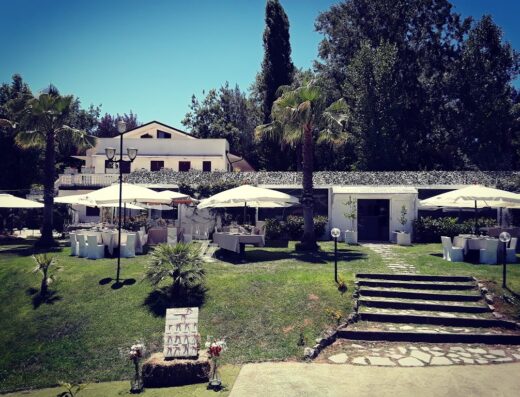 Parco d'Arte Alt Art: Vista esterna di un'elegante location per eventi con tavoli apparecchiati sotto ombrelloni bianchi, prato verde curato e una costruzione bianca sullo sfondo, ideale per matrimoni e ricevimenti.