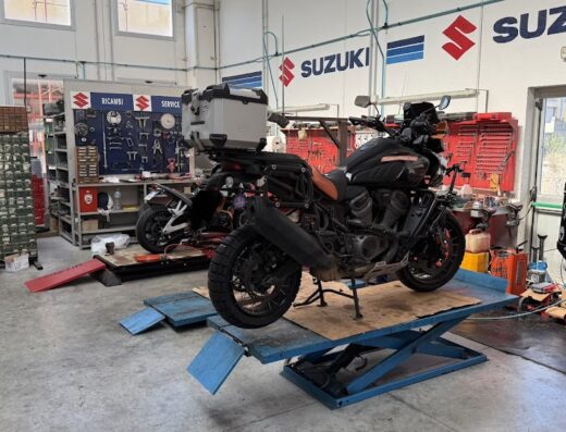 Moto da turismo Harley-Davidson Pan America su piattaforma elevatrice in officina Per Ata Racing di Perria Atanasio, con accessori viaggio e logo Suzuki sullo sfondo.