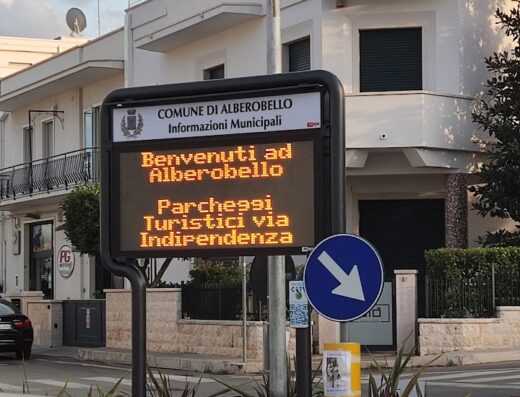 Insegna digitale ad Alberobello, Puglia, che indica "Benvenuti ad Alberobello" e i parcheggi turistici in via Indipendenza. Cartello stradale con freccia blu.