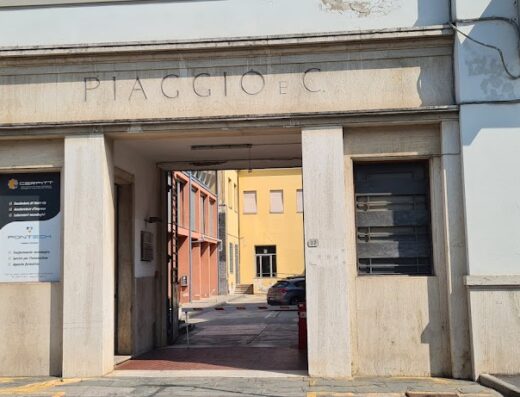 Ingresso principale di Piaggio E C, storico marchio italiano di motocicli, con insegna in pietra e architettura industriale. Vista interna del cortile con edifici e auto parcheggiata.