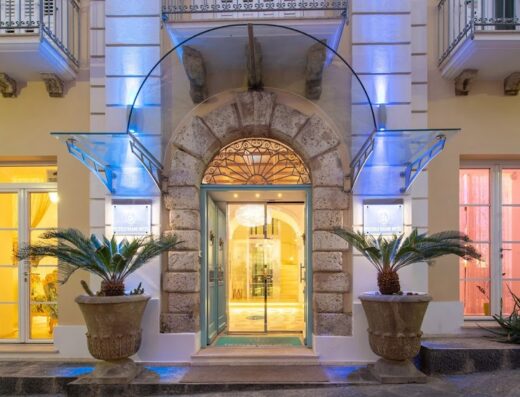 Ingresso principale del Piccolo Grand Hotel Srl, con arco in pietra, vetrata artistica, piante ornamentali in vaso e illuminazione blu decorativa.