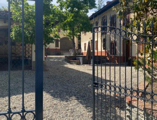 View through wrought iron gates into the courtyard of Piemonte Country House, with gravel driveway, trees, and building facade.