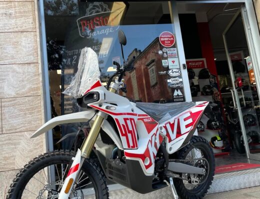 Una moto da rally bianca e rossa parcheggiata di fronte al Pit Stop Garage, un negozio di accessori e servizi per motociclisti, con caschi e ricambi visibili.