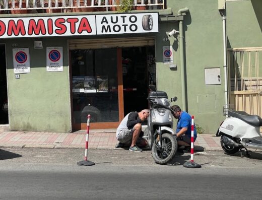 Gommista meccanica pit stop con due meccanici che riparano la gomma di uno scooter di fronte all'officina, con cartello "Gommista Auto Moto".
