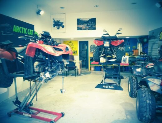 Interno di un'officina PJ Moto con due quad sollevati su piattaforme di sollevamento, accessori moto e attrezzature per riparazioni visibili.