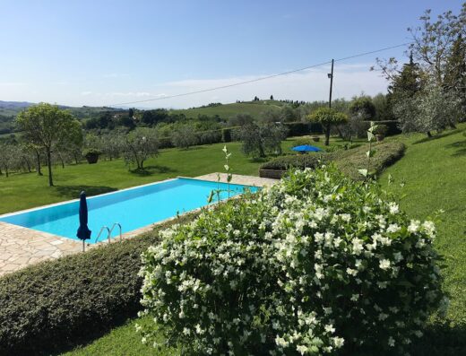 Piscina panoramica al Podere Casanova di Lisa, Toscana, Italia, con ombrellone blu, circondata da giardino curato e colline verdi.