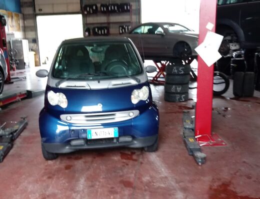 Una Smart blu parcheggiata all'interno di un'officina meccanica con un ponte sollevatore rosso e altre auto sullo sfondo.