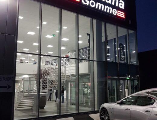 Foto della facciata illuminata di Porrettana Gomme Spa, con vetrine che mostrano l'interno e un'auto di lusso parcheggiata di fronte.