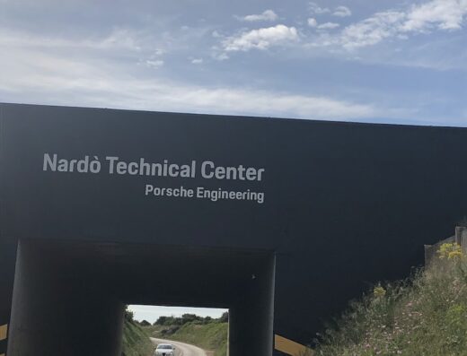 Vista del Nardò Technical Center e Porsche Engineering con auto che percorre la Nardò Ring, un circuito di prova iconico per auto e moto.