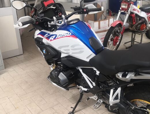 BMW R1250 GS motorcycle in a repair shop, showcasing its blue and white color scheme and off-road tires, with another motorcycle in the background.