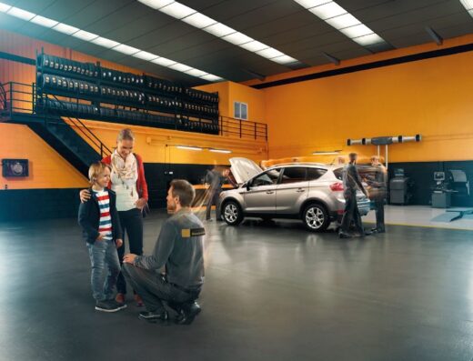 Una famiglia con un bambino parla con un meccanico di BestDrive in un garage con un'auto grigia in riparazione.