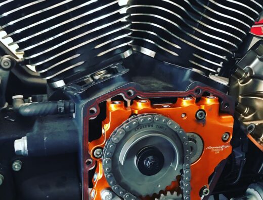 Primo piano dettagliato del motore di una moto, con particolare attenzione alla catena, agli ingranaggi e alla componentistica arancione. Ideale per appassionati di meccanica e riparazione moto.