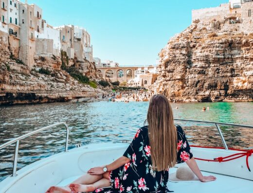 Una donna seduta su una barca a Polignano a Mare, Puglia, con vista sulla spiaggia affollata e sul ponte. Vacanza in barca e servizi turistici.