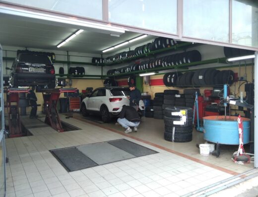 Interno di un'officina Punto Gomme con auto su ponte sollevatore e tecnici al lavoro, scaffali pieni di pneumatici visibili.