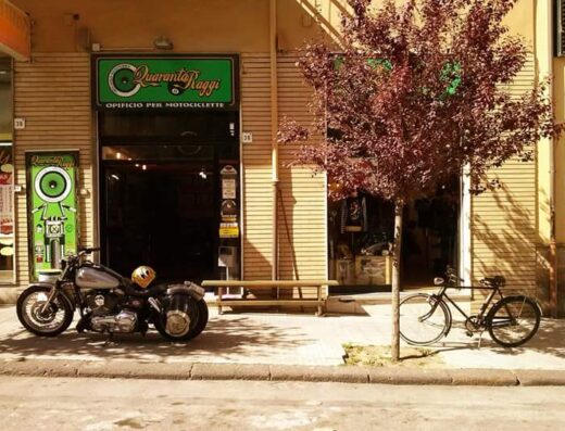 Quaranta Raggi ASD: Motocicletta custom parcheggiata di fronte all'officina per motociclette, con casco giallo sopra. Accanto, una bicicletta classica sotto un albero.