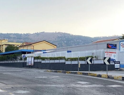 Foto del Quick Washing LAVAUTO dal 1976, situato in una zona urbana con cartelli stradali e sfondo montuoso, che offre servizi di autolavaggio.