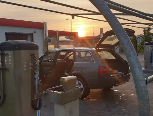 Auto grigia station wagon con portellone aperto in un autolavaggio self-service all'aperto al tramonto. Si vedono aspirapolvere, gettoni e box di lavaggio.