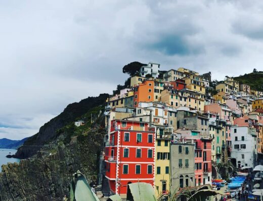 Una vista panoramica di Riomaggiore, Cinque Terre, con case colorate arroccate sulla costa rocciosa. Barche ormeggiate aggiungono colore alla scena marittima.