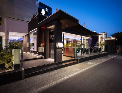 Esterno dell'Hotel Raffaello con il ristorante MOKA, illuminato di sera, che mostra l'architettura moderna e i posti a sedere all'aperto.