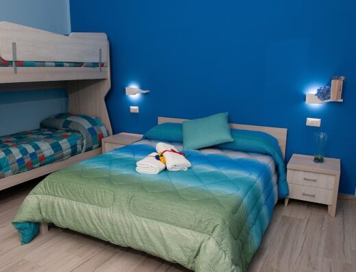 Camera da letto colorata con letto a castello e letto matrimoniale con coperta e cuscini nei toni del blu e del verde, asciugamani bianchi piegati sopra.