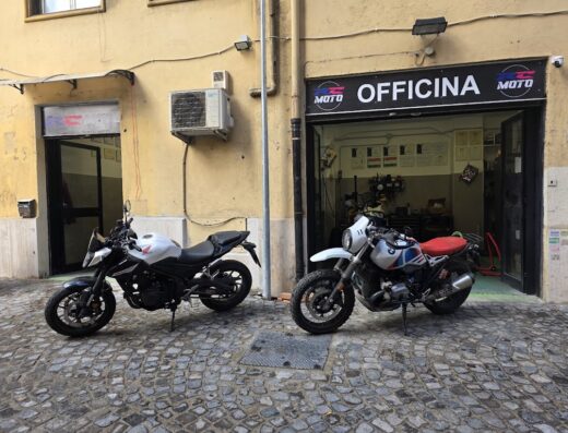 Due motociclette parcheggiate di fronte all'officina RC Moto: una Honda bianca e nera e una BMW bianca con sella rossa. Servizi di riparazione e manutenzione moto.