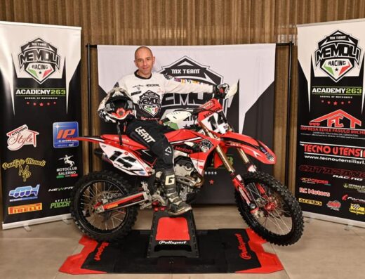 Pilota di motocross professionista posa sulla sua moto Honda numero 14, con attrezzatura Leatt e casco, davanti a banner promozionali di Memoli Racing Academy 263 e sponsor vari, inclusi RC Motors. Ideale per appassionati di motociclismo e gare.