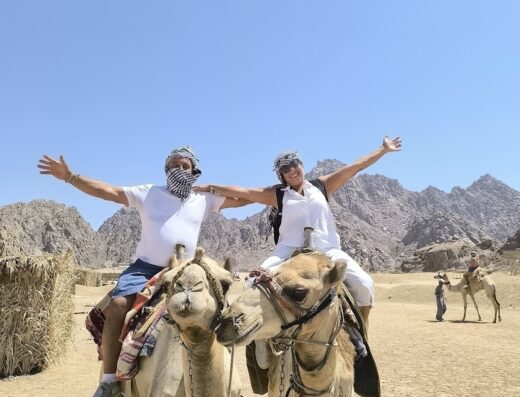 Due turisti sorridenti con turbanti in testa e braccia aperte, seduti su cammelli in una gita turistica nel deserto, offerta da Red Orange Tours and Travel a Palermo.