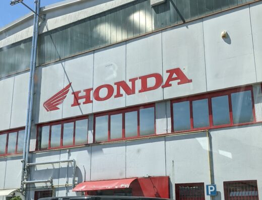 Immagine della facciata di un edificio Honda con furgone nero e auto blu parcheggiate di fronte, sotto un cielo azzurro.