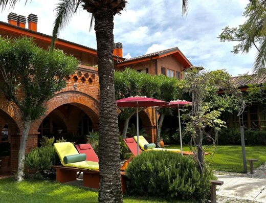 Exterior view of Relais San Damian featuring a lush green lawn, palm tree, lounge chairs with umbrellas, and the building's distinctive orange brick facade.