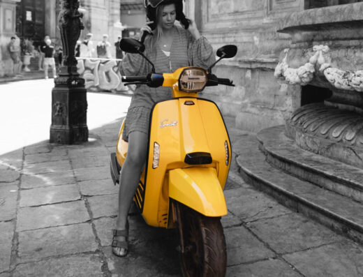 Una donna con casco guida uno scooter giallo in una piazza italiana. Noleggio moto e scooter per esplorare le città.