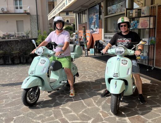 Due turisti in sella a Vespa azzurra di fronte al negozio di noleggio scooter e e-bike a Salò, pronti per un tour sul Lago di Garda.