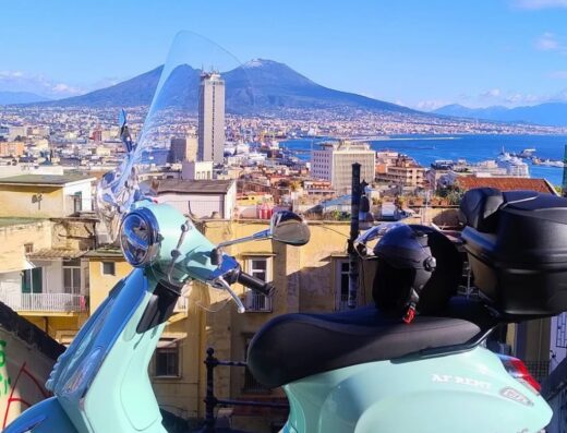 Noleggio scooter Vespa a Napoli: Vista panoramica della città con Vesuvio, scooter turchese con casco e bauletto, AF Rental Napoli.