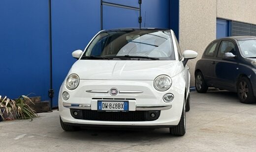Fiat 500 bianca parcheggiata di fronte a una porta blu, con un'altra auto scura visibile sullo sfondo. Noleggio auto e servizi di trasporto a RentalKles.