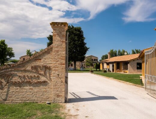 Ingresso di Residence Casale Torrenova Al Conero, con cancello aperto, vialetto, prato curato e edifici tipici sullo sfondo sotto un cielo azzurro.